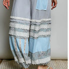 A person wearing loose, wide-leg patchwork pants in light blue and gray with white lace trim, paired with a white lace top and beige sandals, standing against a plain light gray wall.