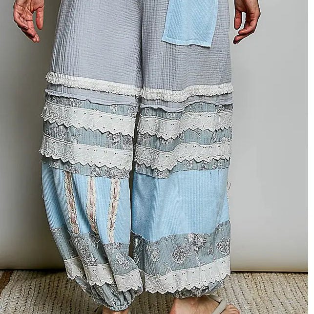A person wearing loose, wide-leg patchwork pants in light blue and gray with white lace trim, paired with a white lace top and beige sandals, standing against a plain light gray wall.