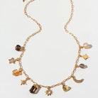 A gold chain necklace with multiple celestial and geometric charms, including stars, moons, planets, and a heart, p;us smoky quartz gemstones displayed on a clean white background.