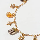 Gold chain charm bracelet featuring star and crystal charms, including a smoky quartz point, faceted amber beads, and clear rhinestone-studded stars, shown on a white background.