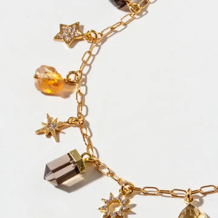 Gold chain charm bracelet featuring star and crystal charms, including a smoky quartz point, faceted amber beads, and clear rhinestone-studded stars, shown on a white background.