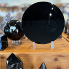 A display of polished black obsidian crystal points and spheres on a wooden shelf, including a large circular black obsidian stone on a clear stand in the background and several faceted black crystal towers in the foreground.