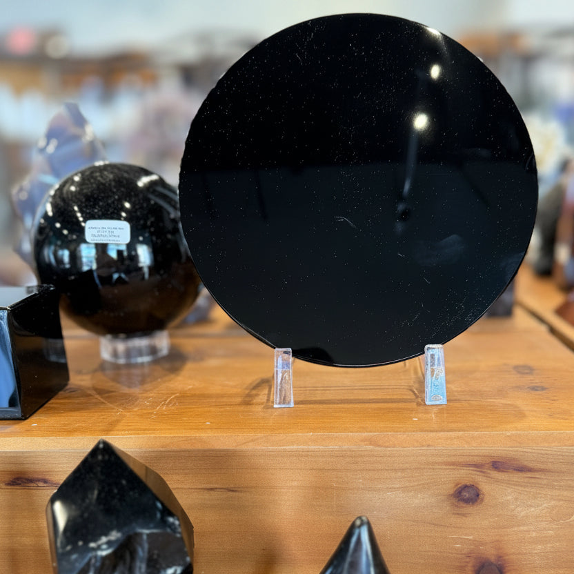A display of polished black obsidian crystal points and spheres on a wooden shelf, including a large circular black obsidian stone on a clear stand in the background and several faceted black crystal towers in the foreground.
