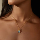 A close-up of a model's neck and upper chest wearing a delicate gold chain necklace with a teardrop-shaped iridescent blue labradorite pendant with gold snake curling around it.