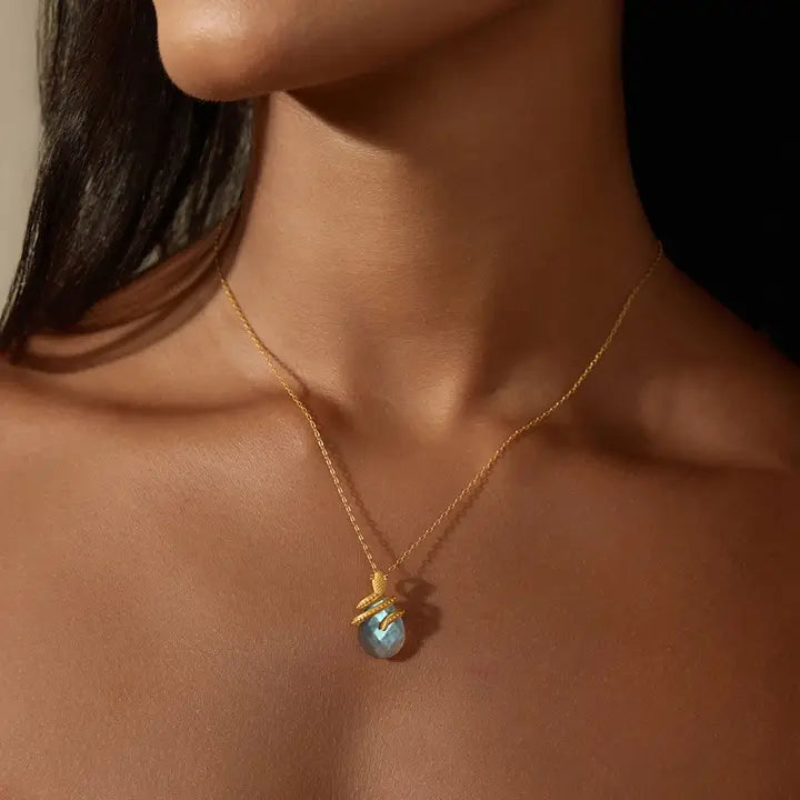 A close-up of a model's neck and upper chest wearing a delicate gold chain necklace with a teardrop-shaped iridescent blue labradorite pendant with gold snake curling around it.
