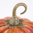 Close-up of a decorative pumpkin with a metallic gold twisted stem and a glossy orange surface with subtle iridescent highlights, set against a plain white background.