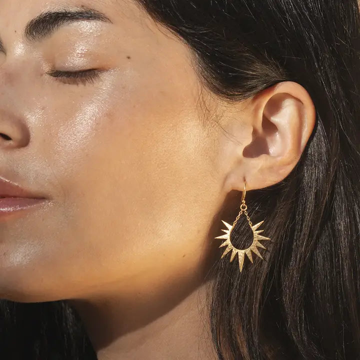 A close-up of a woman's ear wearing a gold sunburst dangle earring with a dark teardrop-shaped inlay, set against warm sunlight and dark hair.
