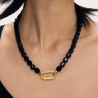 A close-up of a model's neck and chest wearing a black faceted obsidian bead necklace with a textured gold oval clasp.