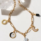 Gold-tone chain necklace with celestial charms including a sun, moon, half moon, star, and black stone accent, displayed on a light surface with a crystal.