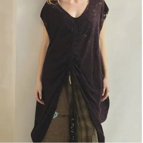 A woman modeling a dark brown sleeveless draped tunic top with a V-neck and gathered front, paired with plaid pants, standing against a neutral beige wall.
