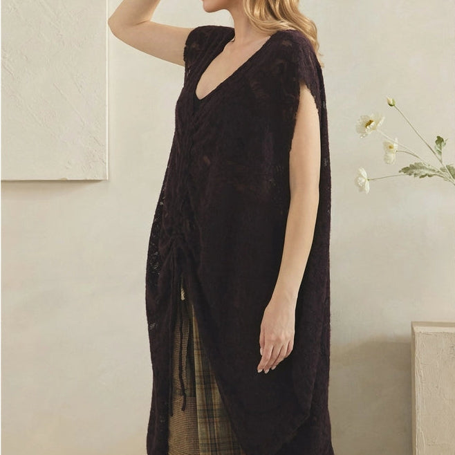 A woman wearing a dark brown sleeveless lace-trimmed tunic dress with a V-neckline, styled over a plaid skirt, standing against a neutral beige wall.