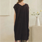 A woman stands with her back to the camera wearing a dark brown sleeveless lace tunic over patchwork wide-leg pants. The background is a minimalist beige room with a white vase of flowers on a stone pedestal.