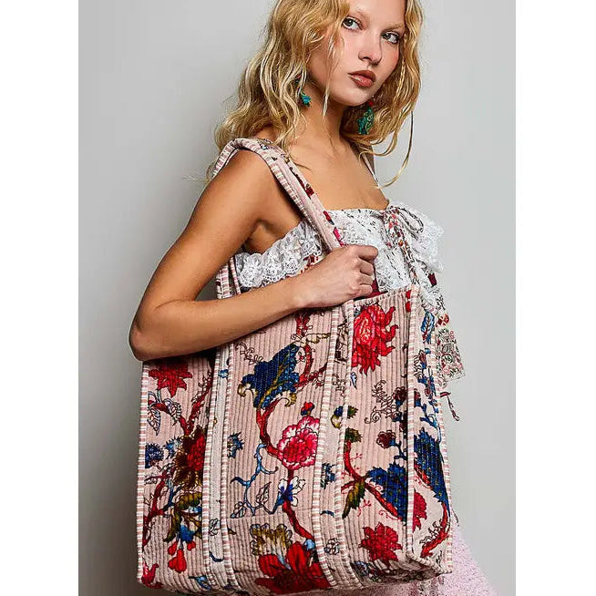A woman models a large, structured tote bag with a pink base and a colorful floral and bird print. The bag features vertical pleated detailing and is shown against a plain light gray studio background.