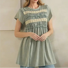 A woman wearing a sage green short-sleeve tunic top with cream horizontal lace stripes and smocked detailing, paired with light blue jeans, standing against a neutral beige wall.