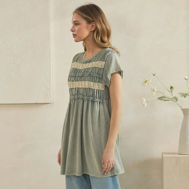 A woman modeling a sage green short-sleeve tunic top with a beige smocked chest panel, paired with wide-leg light blue jeans and beige slide sandals, standing in a minimalist beige room.