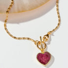 Gold chain necklace with a twisted rope link style and a heart-shaped pink ruby gemstone pendant in a gold bezel setting, displayed on a white surface with a soft beige shell prop.
