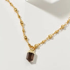 A gold-toned beaded chain necklace with a faceted smoky quartz crystal pendant, displayed on a clean white surface.