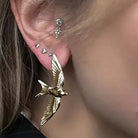 A close-up of a person's ear showcasing a gold-toned bird earring with wings spread, along with several small silver ear piercings.