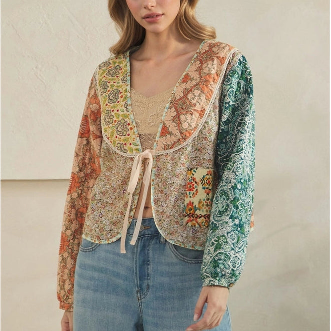 A woman modeling a long-sleeve bohemian-style cropped cardigan with a tie-front detail, featuring a beige floral pattern on the body and sleeves, and a green patterned yoke, paired with light blue jeans against a neutral beige wall.