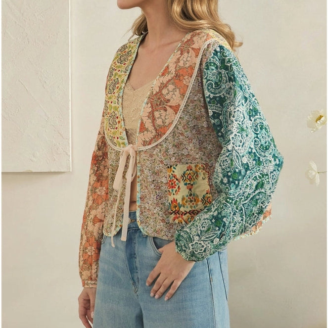 A woman wearing a bohemian-style patchwork vest over a beige top and light blue jeans, standing against a neutral beige wall.