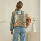 A woman stands with her back to the camera wearing a cropped, long-sleeve patchwork blouse with green, orange, and yellow floral patterns, paired with light blue wide-leg jeans. She is photographed in a minimalist indoor setting with a beige wall and a stone pedestal holding a white vase with flowers.