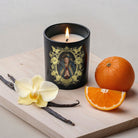 Candle with decorative label on a wooden board with vanilla beans, an orchid, and oranges.