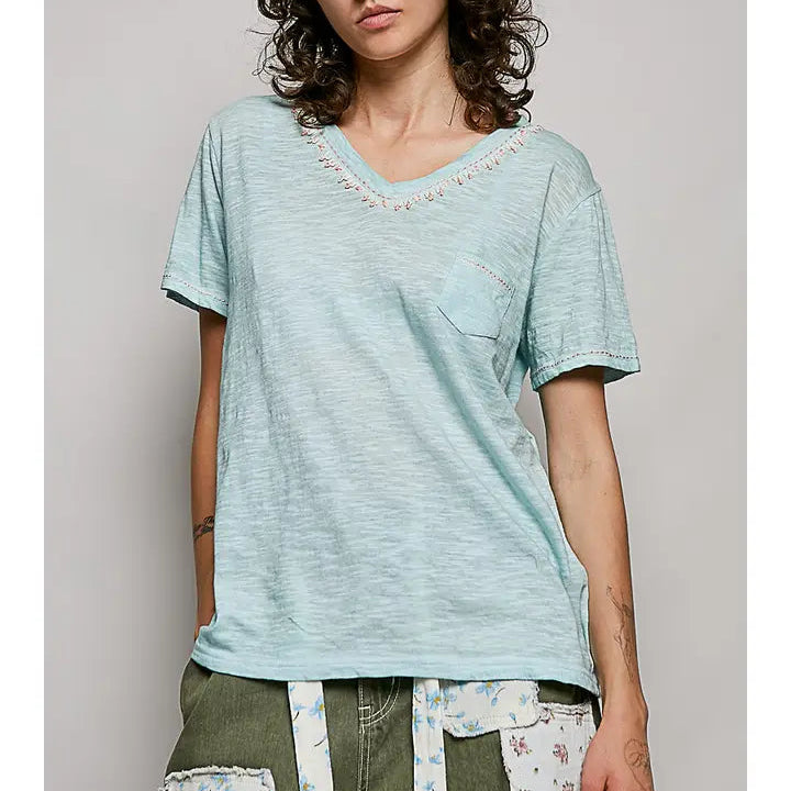 A model wearing a light blue-green V-neck t-shirt with delicate floral embroidery around the neckline, paired with olive green patchwork cargo pants, against a solid light gray background.