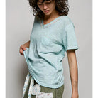 A woman wearing a light mint green V-neck short-sleeve t-shirt with a chest pocket and delicate lace trim, paired with olive green pants and a floral apron, against a neutral gray background.