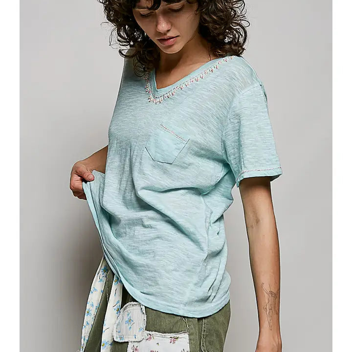 A woman wearing a light mint green V-neck short-sleeve t-shirt with a chest pocket and delicate lace trim, paired with olive green pants and a floral apron, against a neutral gray background.