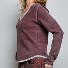 A woman wearing a long-sleeve waffle-knit top in a dusty burgundy color with white lace trim along the V-neck and hem, paired with patterned jeans, against a light gray studio background.