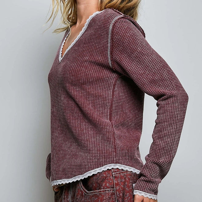 A woman wearing a long-sleeve waffle-knit top in a dusty burgundy color with white lace trim along the V-neck and hem, paired with patterned jeans, against a light gray studio background.