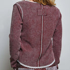 A woman shown from the back wearing a textured, maroon and beige houndstooth-style jacket with white contrast stitching and a lace hem, paired with patterned trousers, against a plain light gray background.