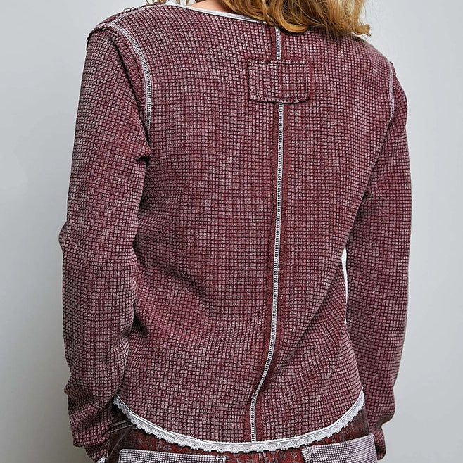 A woman shown from the back wearing a textured, maroon and beige houndstooth-style jacket with white contrast stitching and a lace hem, paired with patterned trousers, against a plain light gray background.