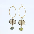 A pair of gold hoop dangle earrings featuring faceted smoky quartz beads and small green labradorite beads, shown against a light blue background.