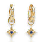 A pair of gold huggie hoop earrings with small clear stones and a dangling starburst charm featuring a blue sapphire gemstone in the center, shown on a white background.