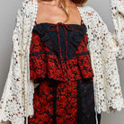 A woman wearing a bohemian-style outfit featuring a red and black floral lace dress with a black lace corset top, a cream crochet cardigan, and a black wide-brim hat, photographed against a neutral gray studio background.