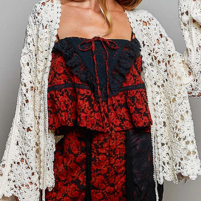 A woman wearing a bohemian-style outfit featuring a red and black floral lace dress with a black lace corset top, a cream crochet cardigan, and a black wide-brim hat, photographed against a neutral gray studio background.