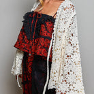 A woman wearing a black wide-brim hat, a red and black floral lace top, matching bottoms, and a cream crochet cardigan, posed against a neutral gray studio background.