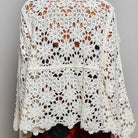 A woman shown from the back wearing a cream-colored, open-knit crochet cardigan with a scalloped hem and wide sleeves, paired with a black wide-brim hat and a red patterned skirt, against a plain gray background.
