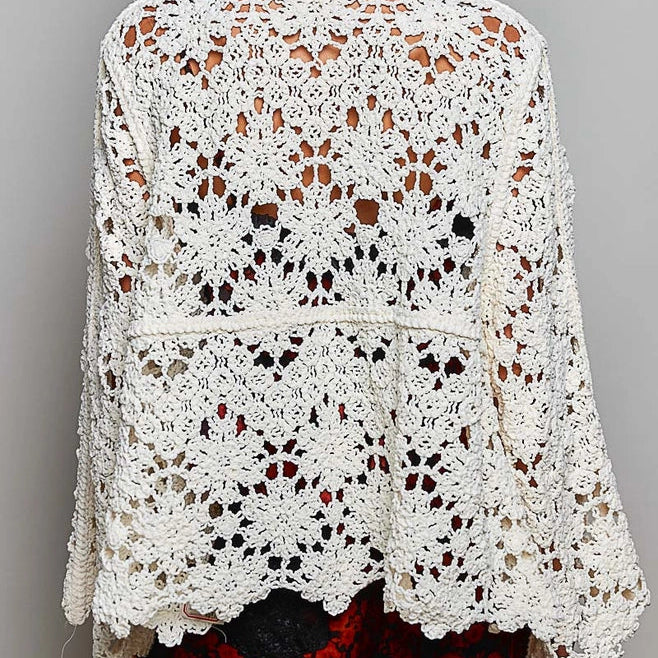 A woman shown from the back wearing a cream-colored, open-knit crochet cardigan with a scalloped hem and wide sleeves, paired with a black wide-brim hat and a red patterned skirt, against a plain gray background.