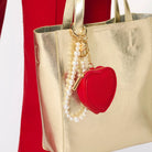 Gold handbag with a red heart-shaped keychain charm and pearl necklace on a white background