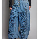 A person wearing high-waisted wide-leg blue denim pants with large black floral embroidery, paired with a dark long-sleeve top and black heeled boots, shown against a plain gray studio background.