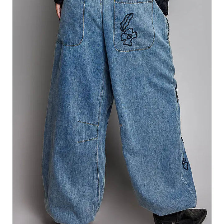 Wide-leg blue denim pants with a drawstring waist, back pockets, and embroidered rabbit motifs on the back pocket and side seam, shown on a model from the back against a gray studio background.