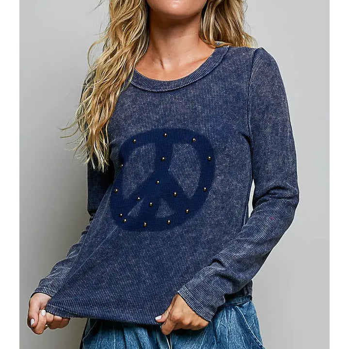 A woman wearing a dark blue long-sleeve t-shirt with a large peace sign graphic on the front, embellished with small gold studs, paired with distressed blue jeans against a plain gray studio background.