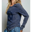 A woman wearing a dark blue long-sleeve ribbed knit top with a large distressed peace sign graphic on the front, embellished with small gold studs, paired with blue jeans against a neutral gray background.