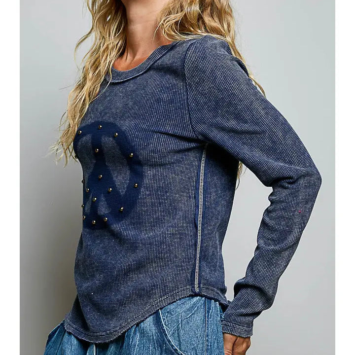 A woman wearing a dark blue long-sleeve ribbed knit top with a large distressed peace sign graphic on the front, embellished with small gold studs, paired with blue jeans against a neutral gray background.