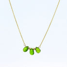 A delicate gold chain necklace featuring three faceted green tourmaline gemstone beads in a row on a light blue background.