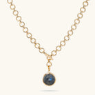 A gold chain necklace with a toggle clasp and a round bezel-set dark labradorite stone pendant, shown on a light cream background.