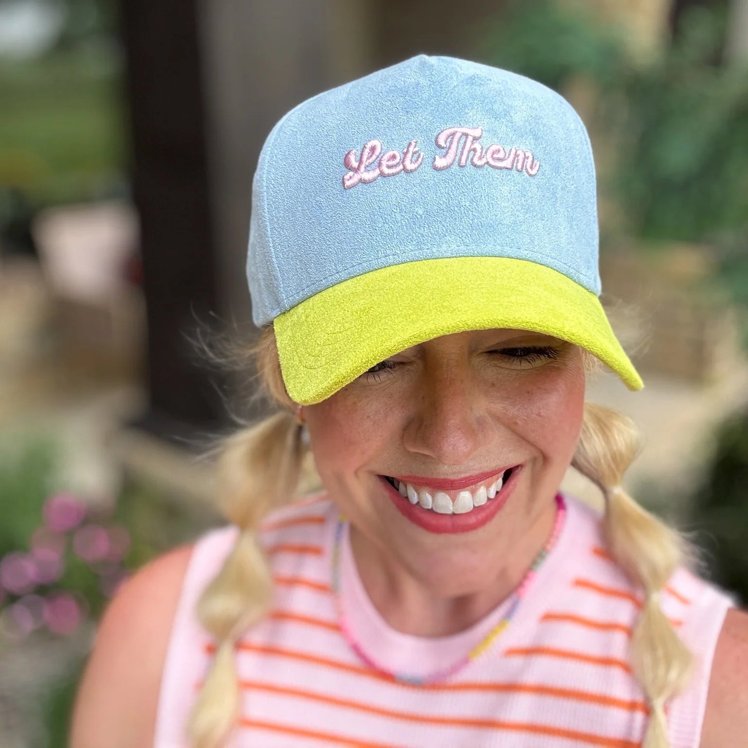Woman wearing a blue and yellow cap with 'Let Them' text, smiling outdoors.