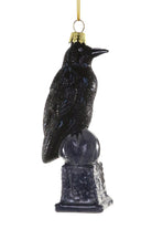 A black raven Christmas ornament with a glittery finish, perched on a dark pedestal base with a gold hanging loop at the top, shown against a plain white background.